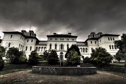 Gothic mansion with dramatic clouds overhead and a fountain in front.
