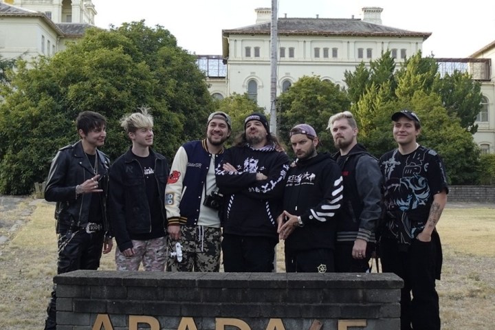 Group of seven people standing behind a stone sign reading 'ARADAKE' with a building in the background.